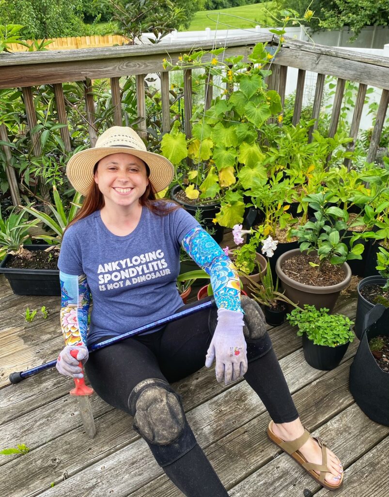 A women sitting on a deck in front of a group of potted plants. She is wearing a straw hat, tshirt that reads "ankylosing spondylitis is not a dinosaur," and holding a cane.